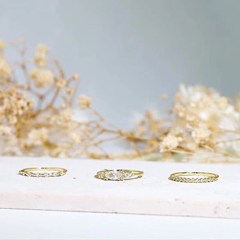 Set of three delicate stackable gold rings with cubic zirconia stones on white surface with blurred floral background